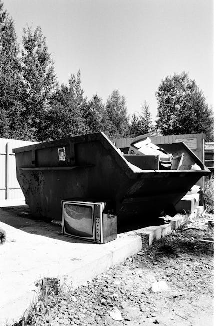 A lineup of four large white wheelie bins with closed lids, positioned on a paved street near a metal waste container and a black, graffiti-style graffiti, inside a property or loading area, with tree